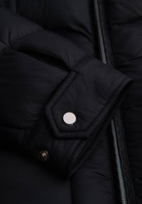 Gros plan d’une manche de veste matelassée noire avec un bouton-pression argenté et une fermeture éclair argentée visible le long de l’ouverture frontale.