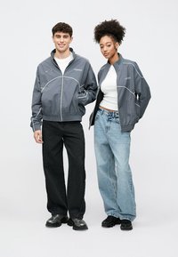 Grå vindjakker med hvite detaljer og glidelås foran, båret av to personer. Den mannlige modellen har på svarte bukser, den kvinnelige har lyse jeans.