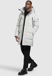 Világosszürke pufi kabát kapucnival, elől cipzárral, két oldalsó zsebbel és logó címkével. A modellen fekete nadrág és vastag fekete csizma van.