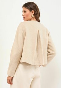 Vrouw in een beige lange mouwen cropped jas met een rugplooi, gecombineerd met hoog-getailleerde lichte crèmekleurige broek, zijdelings kijkend.