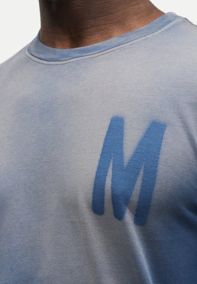 Κοντινό πλάνο ατόμου που φοράει γαλάζιο πουκάμισο με ένα μεγάλο σκούρο μπλε γράμμα "M" τυπωμένο στο στήθος.