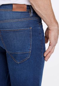 Jeans de mezclilla en azul oscuro con una textura suave. Las características incluyen un parche de cuero marrón con "JACK'S", costuras naranjas en contraste y un bolsillo trasero.