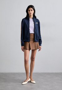 Navy athletic jacket with light stripes, light purple tee with logo, brown pleated skirt, and cream low-heeled shoes. Smooth textures.