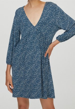Mujer con vestido azul con estampado floral pequeño en blanco, escote en V y mangas largas, de pie con una mano en la cintura y otra al lado.