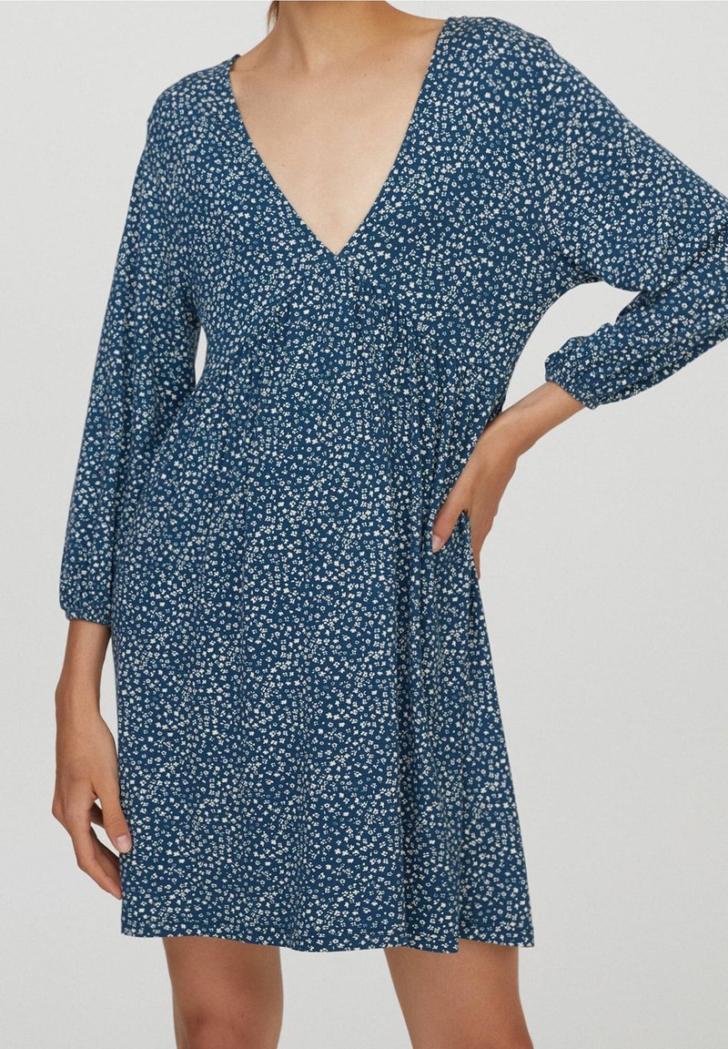 Femme portant une robe bleue à petit imprimé floral blanc, col en V et manches longues, debout avec une main sur la taille et l’autre le long du corps.