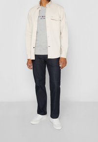 Camisa beige de manga larga sobre una camiseta gris, combinada con jeans azul oscuro y zapatillas blancas. Detalle notable en el bolsillo de la camisa.