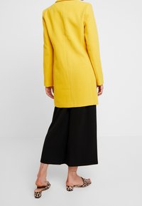 Manteau en laine jaune au design structuré et aux longues manches, associé à un pantalon large noir et des sandales à semelle plate à imprimé léopard.