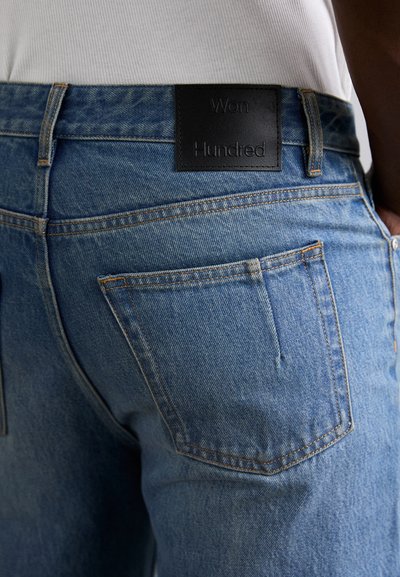 Jeans de mezclilla azul con una etiqueta de cintura de cuero negro en la que está grabado "Won Hundred". La tela tiene un acabado texturizado clásico.