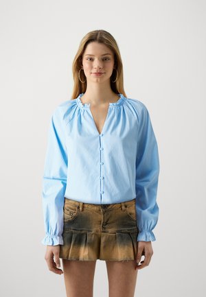 Lys blå bluse med en samlet halsudskæring, lange ærmer og knaplukning, parret med en falmet brun denimnederdel med en friturekant.
