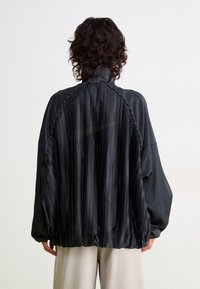 Black satin jacket featuring a pleated back design, high collar, and studded accents along the shoulders. Relaxed fit with long sleeves.