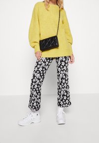 Osoba oblečená v žlutém pleteném svetru, černobílých květinových kalhotách, bílých vysokých teniskách a nese černou prošívanou kabelku p�řes rameno.