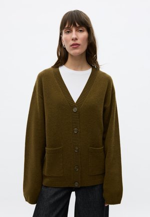 Femme portant un cardigan boutonné vert olive par-dessus une chemise blanche et un pantalon foncé, debout devant un fond clair uni.