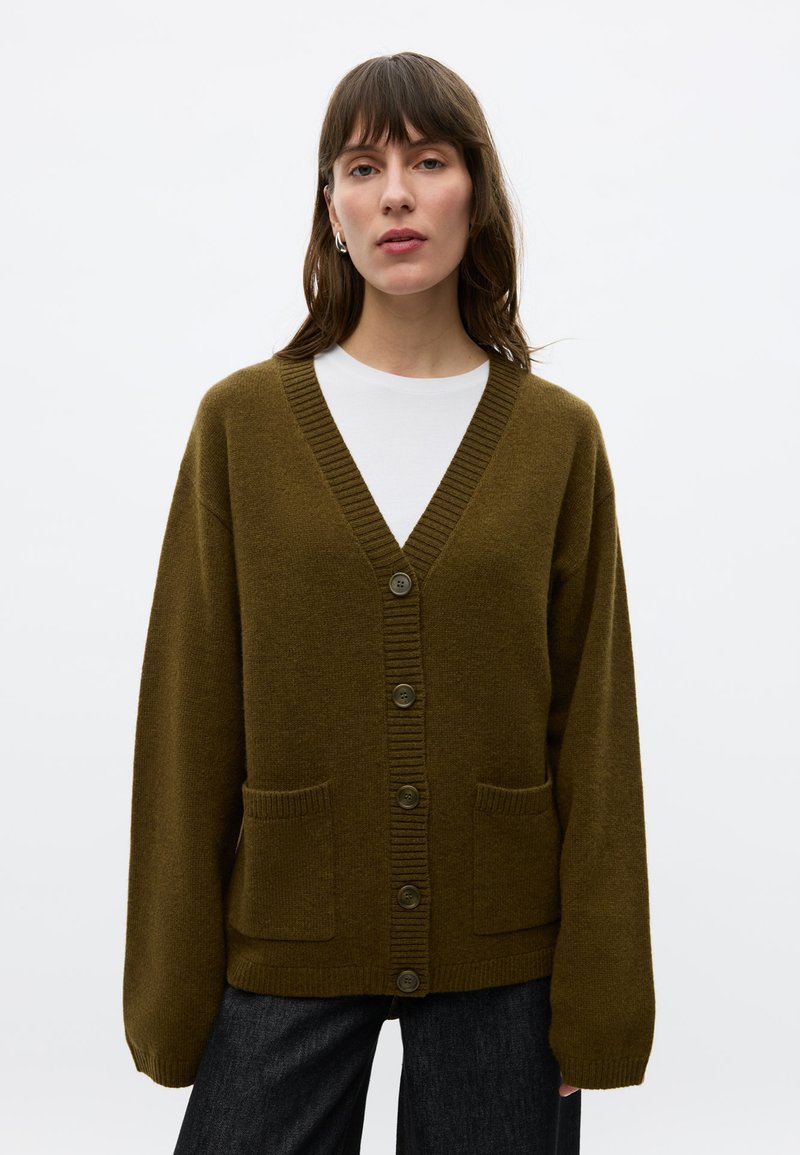 Femme portant un cardigan boutonné vert olive par-dessus une chemise blanche et un pantalon foncé, debout devant un fond clair uni.