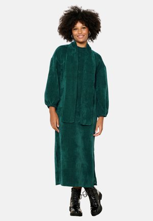 Abito di corda color teal con maniche lunghe e una sciarpa coordinata, caratterizzato da una vestibilità oversize e spacchi laterali, abbinato a stivali neri con lacci.