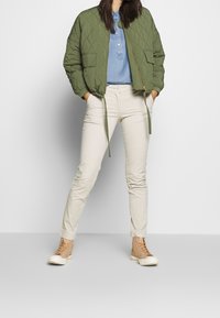 Quilted green bomber jacket, light blue button-up, beige slim-fit pants, and tan high-top sneakers. Textured material and visible seams.