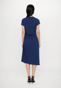 Mörkblå knälång klänning med korta ärmar, med en åtsamlad rygg och en asymmetrisk fåll. Tyget verkar mjukt med en slät textur.