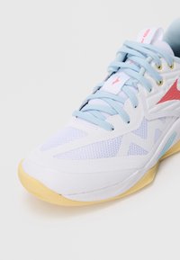 Zapatilla deportiva blanca con cordones azules, paneles de malla y acentos rojos. Presenta una suela texturizada y un diseño elegante con una base amarilla.