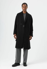 Schwarzer Wollmantel mit Reverskragen, einem einzelnen Knopf und silbernem Schnallendetail, getragen über einem grauen Pullover und grauen Hosen.