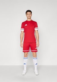 Red sports uniform featuring a short-sleeve shirt and shorts with white stripes, paired with white socks accented by blue stripes and logo.