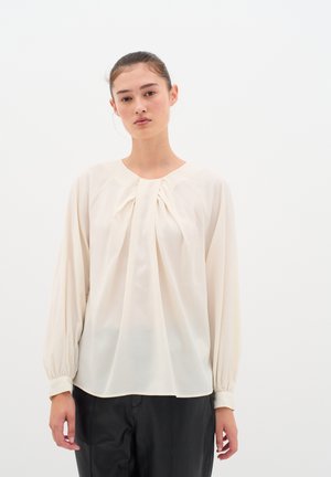 Blusa de color crema con mangas largas y abullonadas y detalle fruncido en la parte delantera. Fabricada en un tejido ligero con una textura suave.