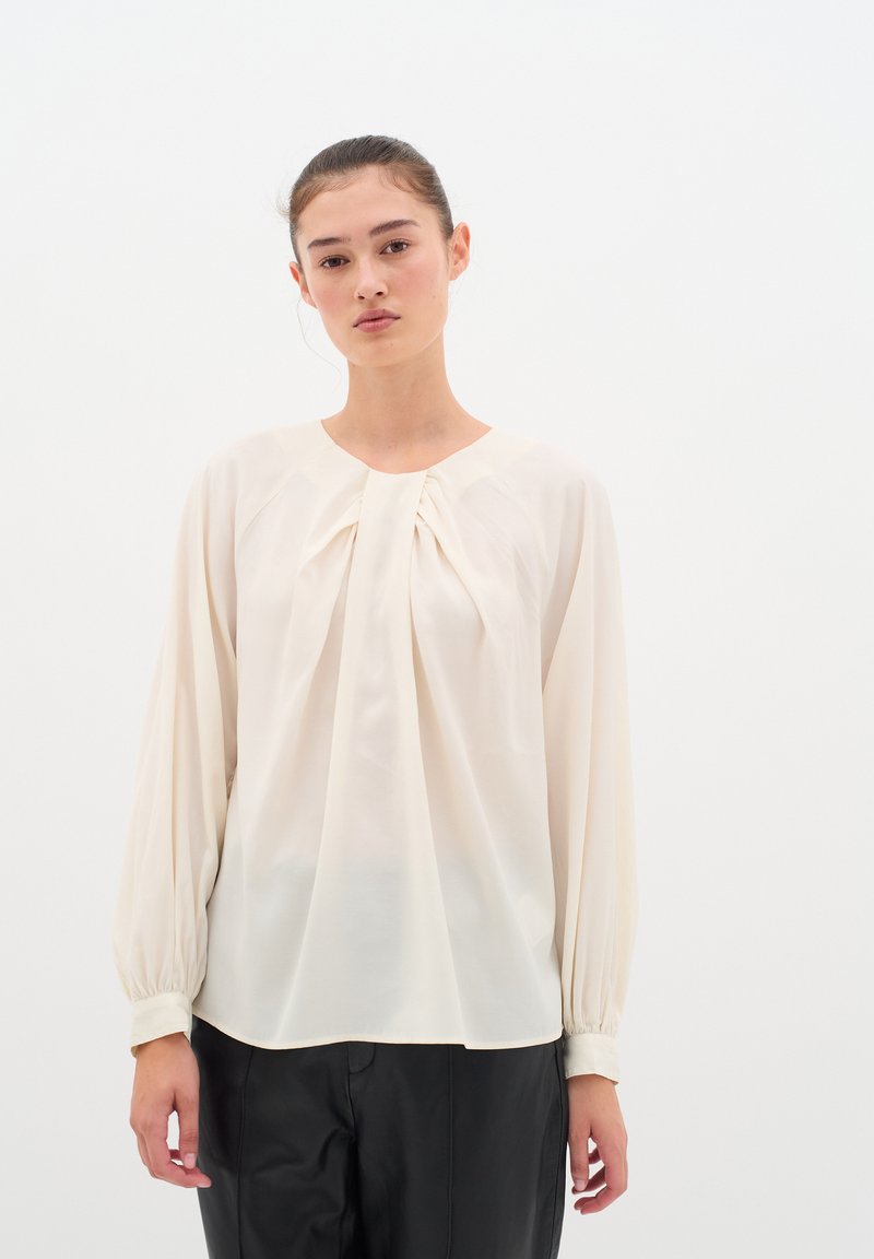 Blusa color crema con maniche lunghe e ampie e dettagli arricciati sul davanti. Realizzata in tessuto leggero con una texture liscia.