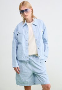 Giacca a righe blu chiaro con dettagli in argento e due tasche frontali, abbinata a pantaloni corti coordinati. Il modello indossa una maglietta bianca e occhiali da sole.