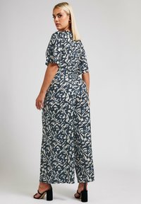 Jumpsuit batik bunga berwarna navy dan krim, menampilkan kaki lebar, lengan pendek, dan pinggang yang pas. Sandal hak tinggi hitam melengkapi penampilan.