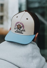 Trucker-Cap mit einem beigen Vorderteil, blauen Schirm und braunem Netzrücken. Der bestickte Entwurf enthält ein Keks-Maskottchen und bunte Schrift.