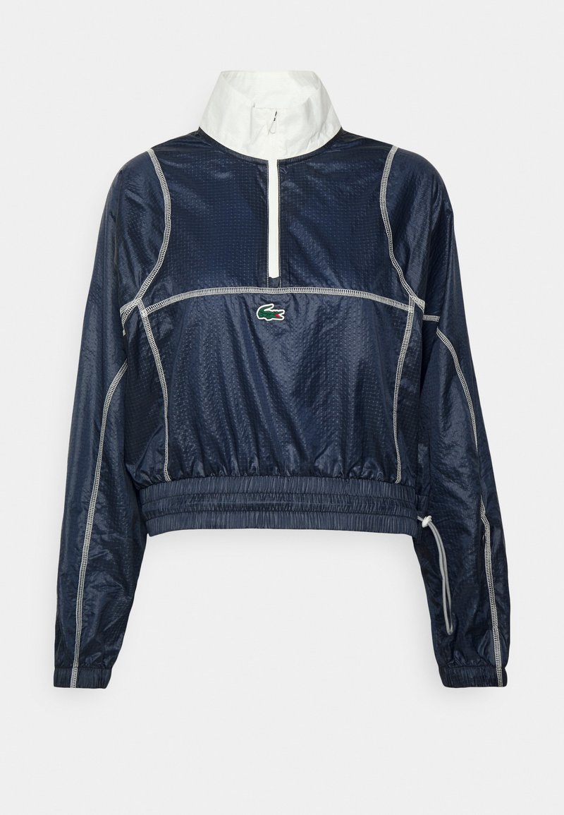 Veste coupe-vent bleu marine avec col blanc, devant zippé à mi-hauteur et ourlet élastique. Présente un logo vert et des accents de couture réfléchissants.