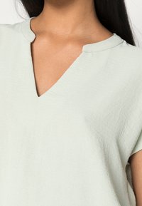 Top corto a maniche corte verde chiaro con scollo a V, realizzato in tessuto testurizzato, caratterizzato da una vestibilità rilassata e dettagli di cucitura sottili.