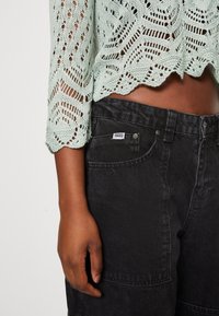 Dunkelhäutige Person trägt schwarze Jeans mit "RAGGED"-Etikett und einen mintgrünen gehäkelten, kurz geschnittenen Pullover mit gewelltem Saum und Ärmeln.