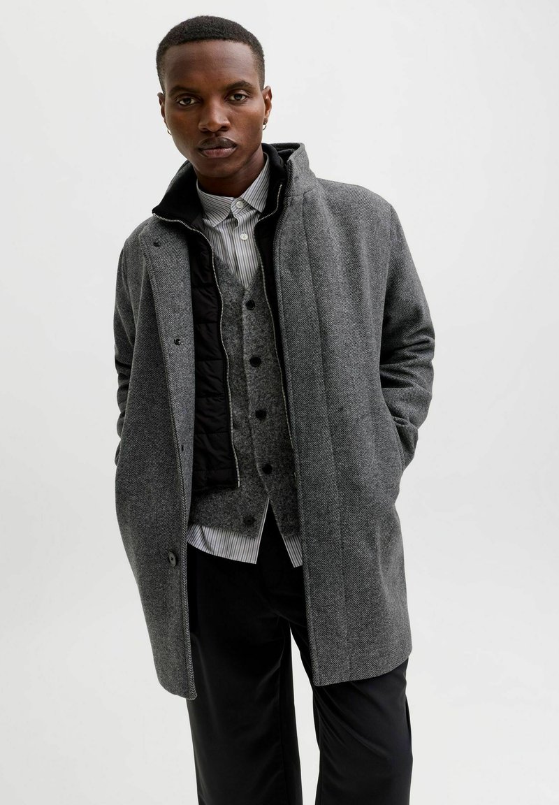 Grijze wollen blend overjas met hoge kraag, gelaagd over een grijze vest en gestreefd shirt. Bevat knopen en heeft een soepele textuur.