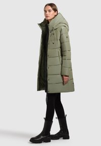 Olīvzaļš pūkains mētelis ar kapuci, ar rāvējslēdzēju priekšpusē, ar auklu regulēšanu un sānu kabatām. Apvienots ar melniem legingiem un potīšu zābakiem.