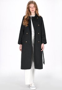 Zwarte trenchcoat met dubbele rij knopen, bronzen knopen, zijzakken en een riem in de taille. Gedragen over een witte outfit, met lange mouwen.
