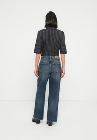 Femme portant une chemise à rayures noires à manches mi-longues, un jean large bleu et des bottines à talons noires, debout face à un mur blanc.