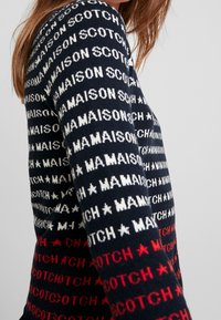 Sweter dziany w granatowym kolorze z powtarzającymi się białymi i czerwonymi wzorami logo. Mankiety z prążkowanego ściągacza i miękka faktura. Dopasowany krój.