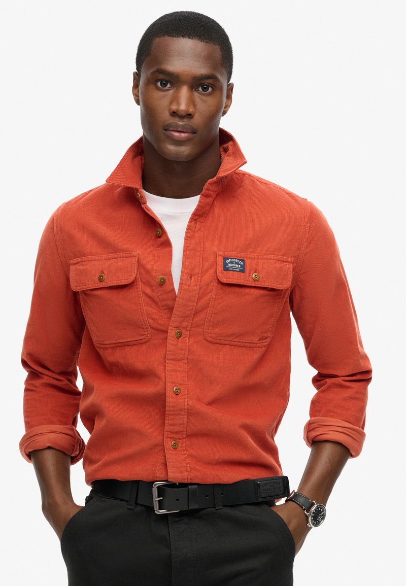 Oranje corduroy shirt met twee borstzakken, met knopen bij de manchetten en een kraag, gedragen over een wit T-shirt en gecombineerd met een zwarte riem.