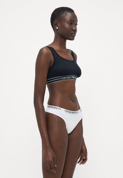Brassière en coton noir avec un bandeau logo, associée à un slip en coton blanc de style bikini, présentant une ceinture logo. Textures lisses, design simple.