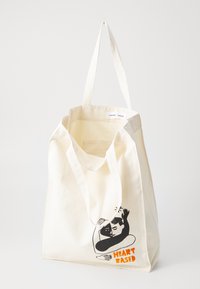 Canvas tote bag in off-white with black illustration of a figure and orange text "HEART BASED". Two shoulder-length handles. Simple design.