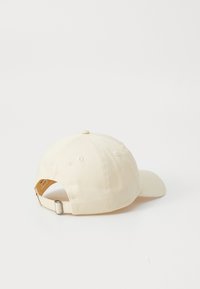 Caps laget av lett stoff, i kremfarge, med en buet skjerm, justerbar metallspenne bak, og sydd øyer for ventilasjon.