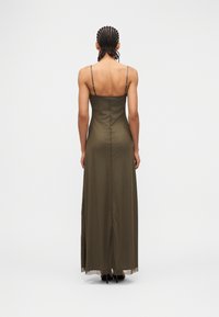 Sieviete ar pītām matu tasiņām, tērpusies garā olīvu zaļā bezroku kleitā un melnos augstpapēžu apavos, stāvot pret vienkāršu baltu fona sienu.