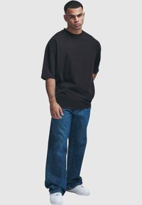 Černé oversized tričko s krátkými rukávy, kombinované s volnými modrými džíny a bílými teniskami. Hladká tkanina s uvolněným střihem.