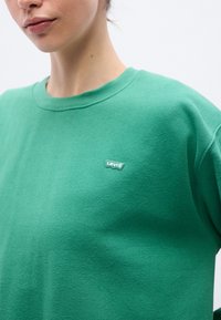 Sweat-shirt en coton vert avec un col rond, arborant un petit patch logo Levi's sur la poitrine. Texture douce, coupe décontractée et couleur unie.