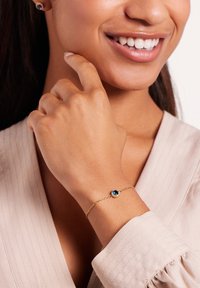 Gouden armband met een blauwe edelsteen, voorzien van een delicate schakels ontwerp. Draagt op een hand, met een textuur, lichtgekleurde kleding.