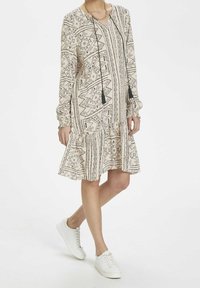 Långärmad klänning i beige med svarta geometriska mönster, flätad volang i nederkant och V-ringning. Stylar med vita sneakers och ett halsband.