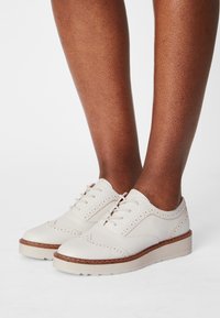Chaussures à lacets en cuir blanc avec une surface texturée, des détails brogues, et une semelle de couleur claire avec une légère élévation.