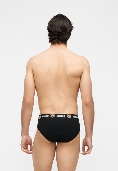Άνδρας όρθιος με την πλάτη στην κάμερα, φοράει μαύρα εσώρουχα με κορδόνι με σχέδια αρκουδάκια και το κείμενο «MOSCHINO».