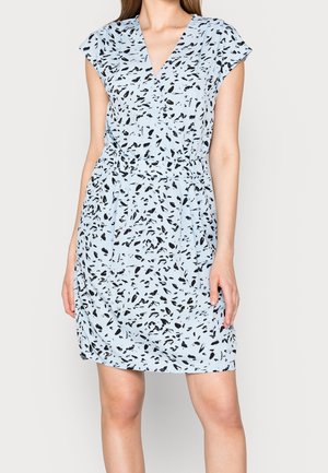 Frau, die ein hellblaues Kleid mit kurzen Ärmeln, schwarzen abstrakten Flecken und V-Ausschnitt trägt, vor einem schlichten hellen Hintergrund steht.