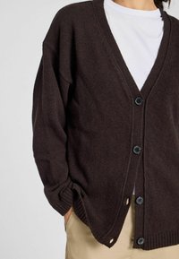 Donkerbruin gebreid vest met een V-hals, geribbelde manchetten en vijf zwarte knopen aan de voorkant. Heeft zijslits voor extra comfort.