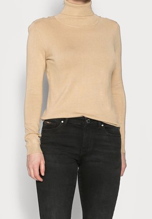 Pull à col roulé beige à manches longues associé à un jean noir taille haute, visible de la moitié du torse jusqu'aux hanches.